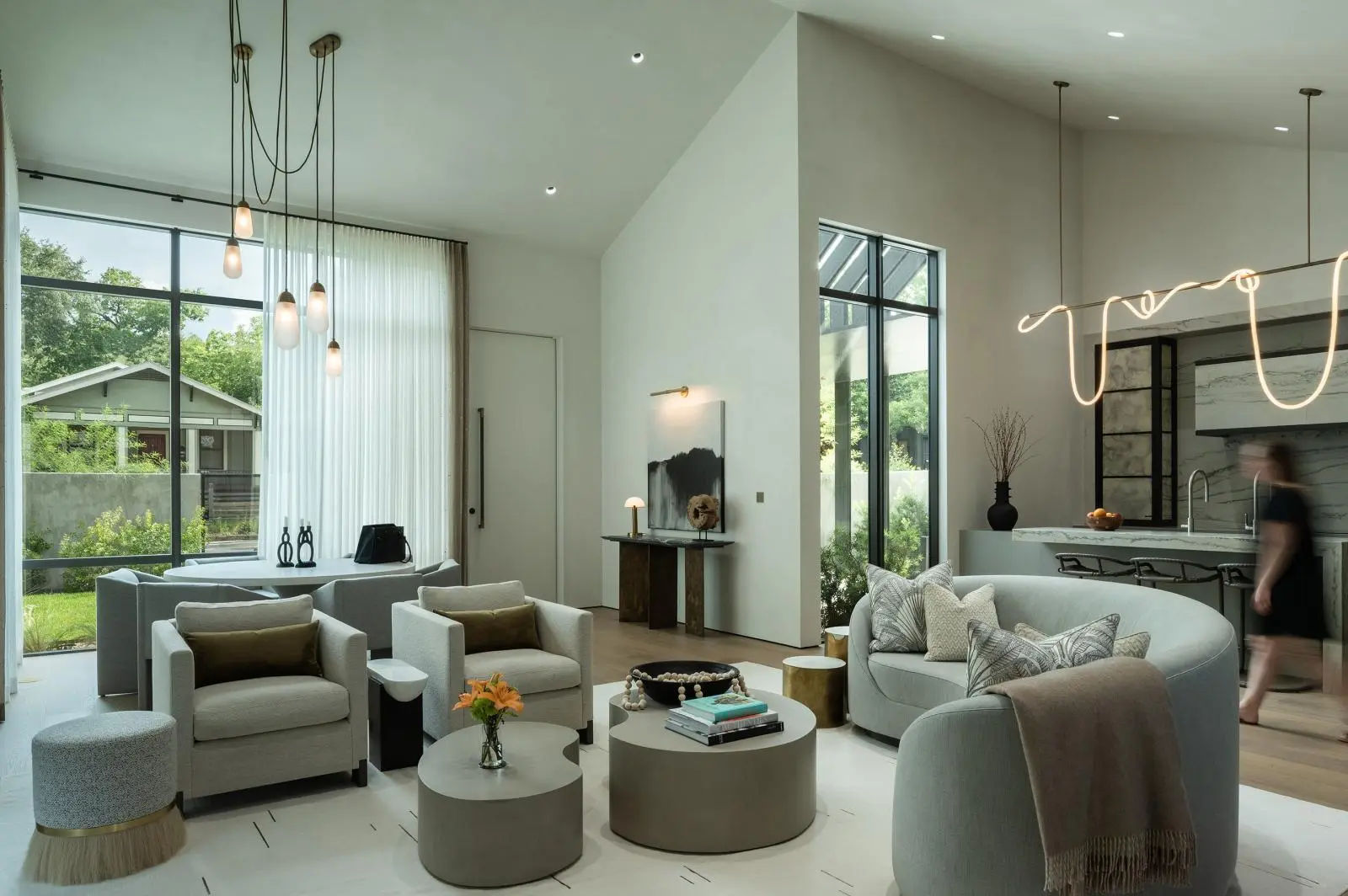 Modern open-plan living room with gray seating, circular coffee tables, and floor-to-ceiling windows letting in natural light; pendant lights hang from the ceiling and sheer curtains frame the view of a garden outside.