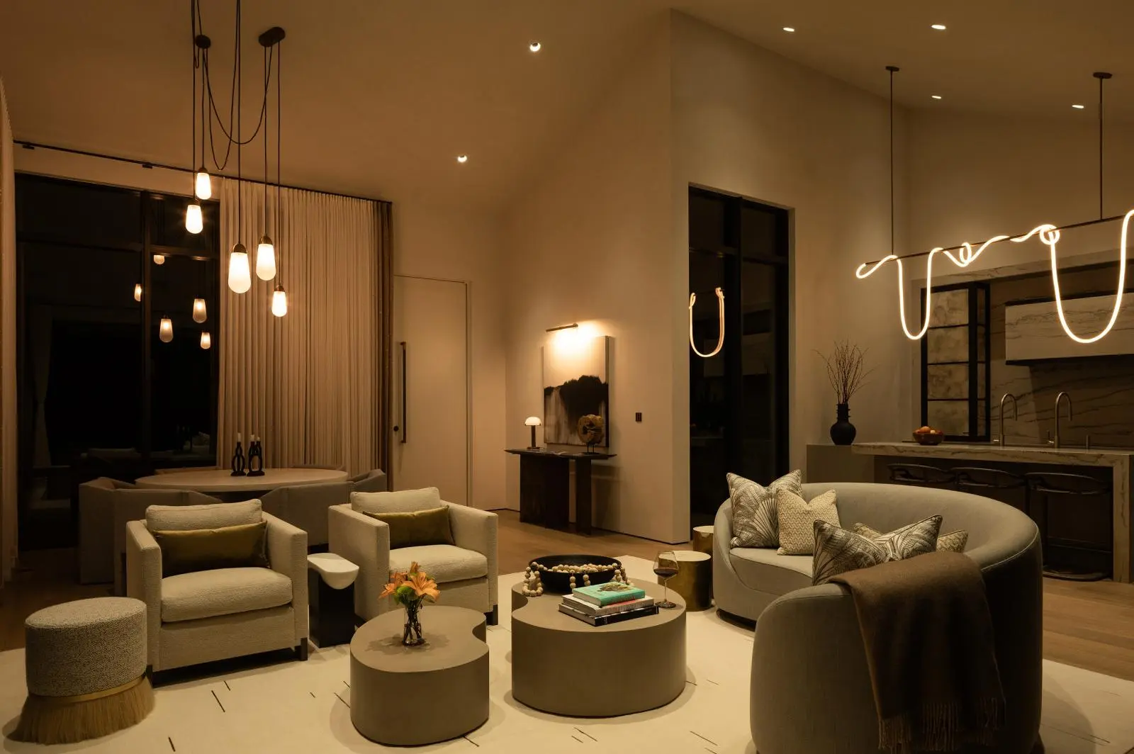 Warm, contemporary living room with beige seating, round coffee tables, and warm pendant lights; open-plan kitchen in background with a glowing light installation above.