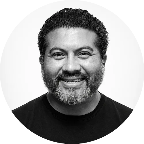 ERIC Head-and-shoulders portrait of a smiling man with dark hair and a gray beard, wearing a black shirt against a light background.