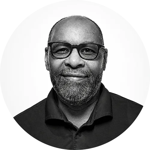 RUDY Portrait of a smiling Black man with glasses and a gray beard, wearing a dark polo shirt, facing the camera.