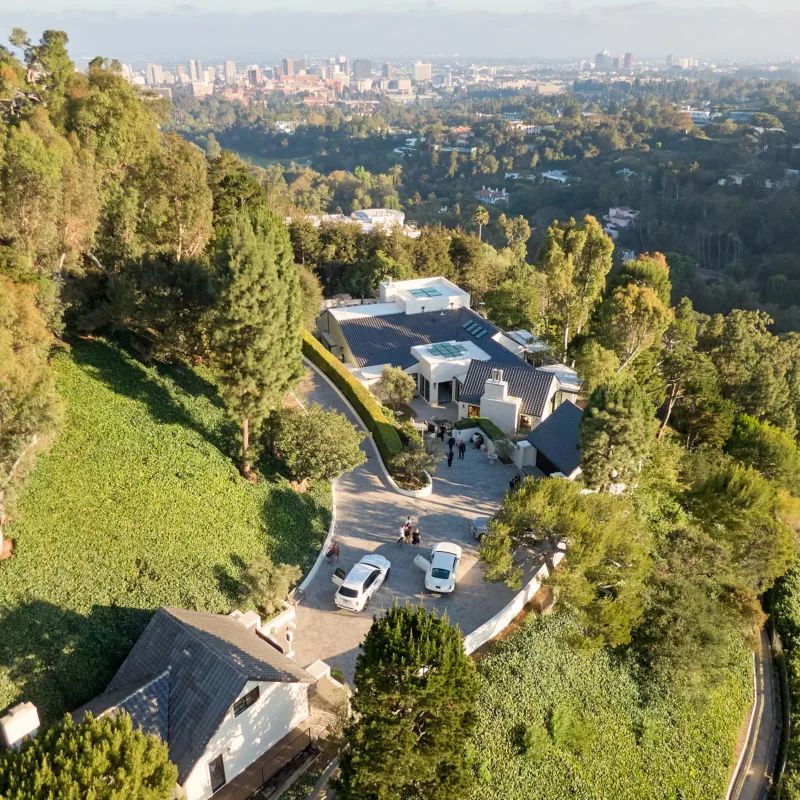 Aerial view of a hillside neighborhood with modern houses, a curved road, and a distant city skyline.