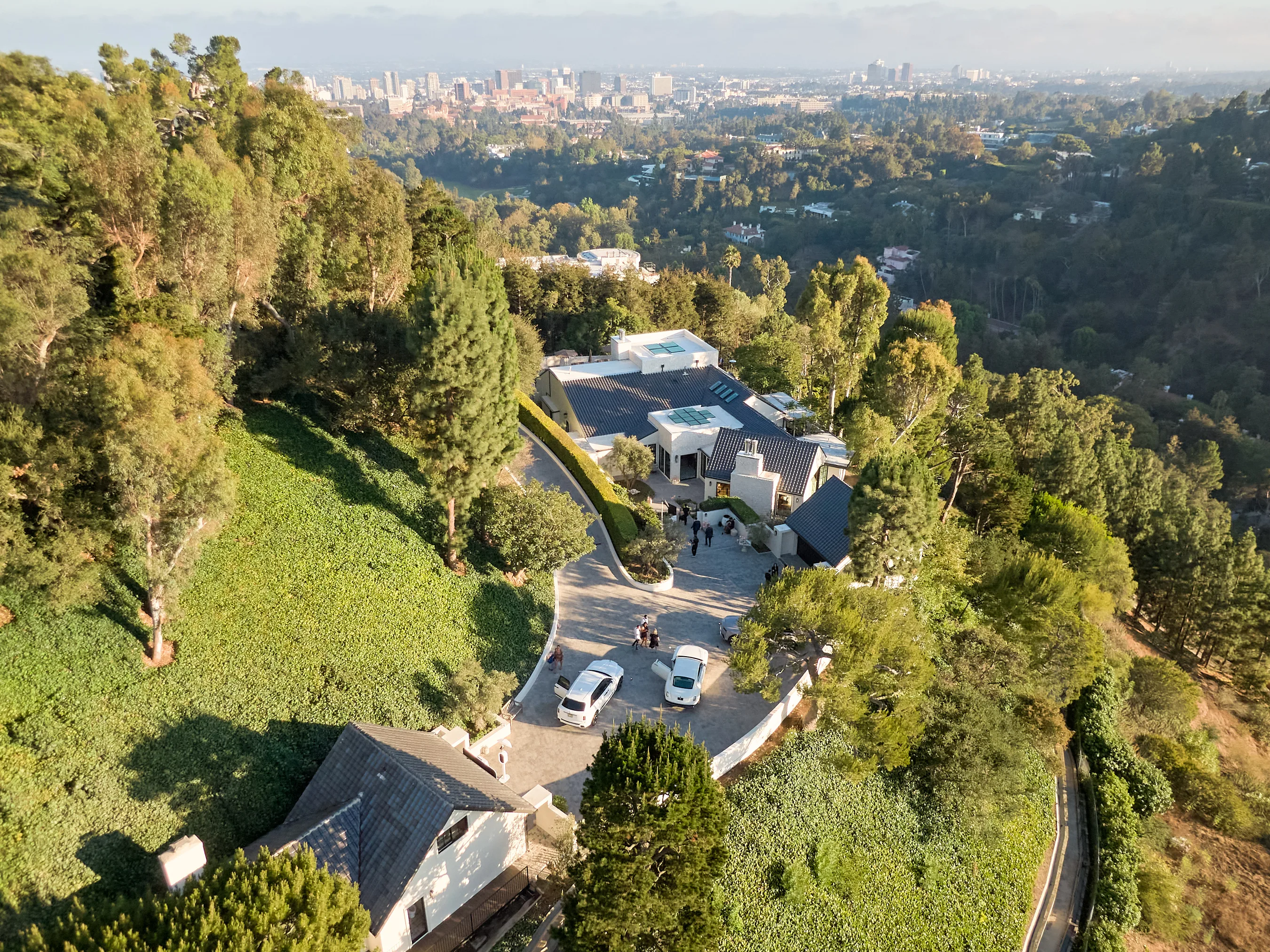 Aerial view of a hillside neighborhood with modern houses, a curved road, and a distant city skyline.