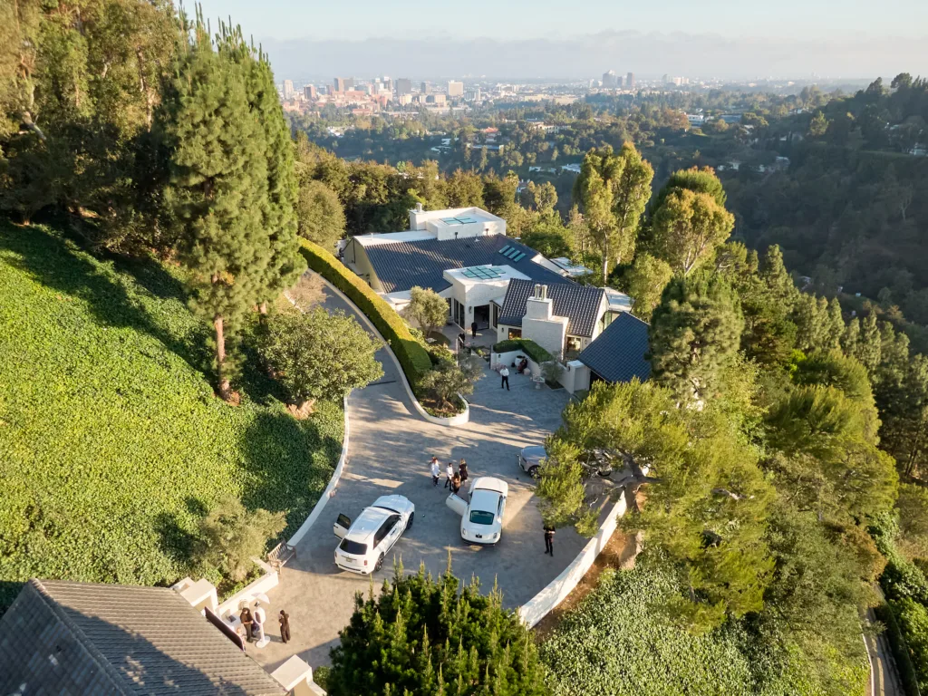 Aerial view of a hillside residential area with modern white houses, a curved driveway, and several parked cars amid lush greenery, overlooking a distant city skyline.