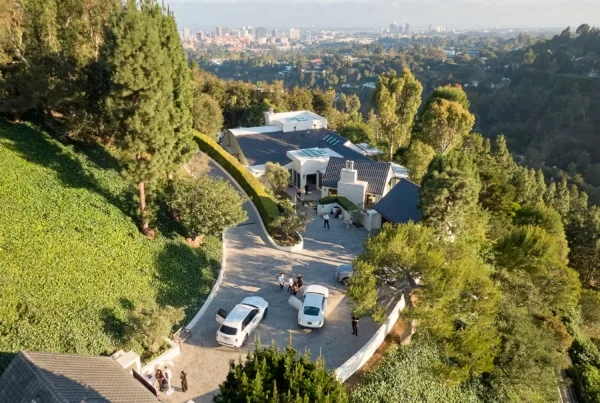 Aerial view of a hillside residential area with modern white houses, a curved driveway, and several parked cars amid lush greenery, overlooking a distant city skyline.