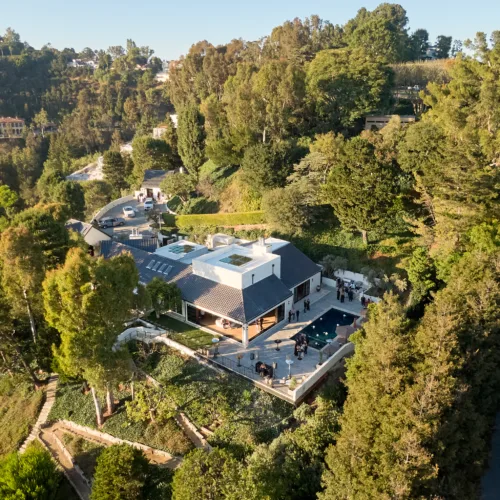 Aerial view of a modern hillside house with a rectangular pool and outdoor seating, surrounded by dense trees and a group of people on the patio.
