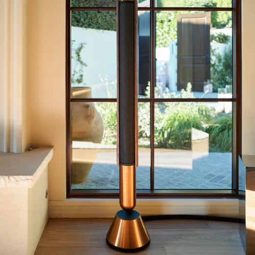 Beolab 28 floor-standing speaker with a black mesh grille beside a large window overlooking a garden area.