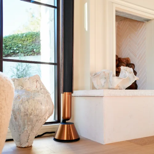 B&O Beolab 28 standing by a window, on a light wood counter with decorative stone pieces nearby.