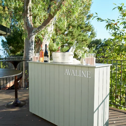 Outdoor bar cart with Avaline branding, bottles and an ice bucket on a wooden deck, with lush trees in the background.