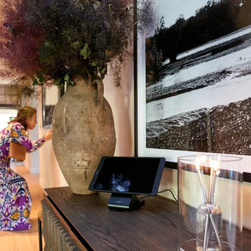 Vase with dried purple flowers on a dark wooden console, with a tablet on a stand and a glass candle holder nearby; a blurred person in colorful clothing is in the background.