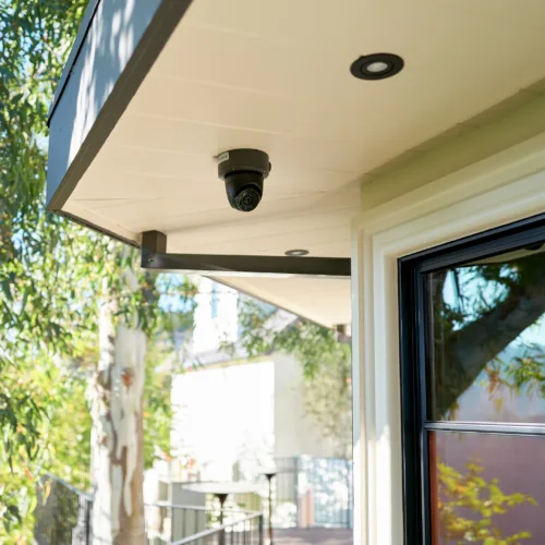 Outdoor security camera mounted under a beige awning beside a window with trees in the background.
