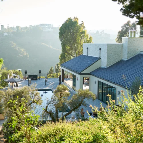 Modern hillside house with a blue tiled roof, stone chimney, and surrounding trees; patio with umbrellas in the foreground.
