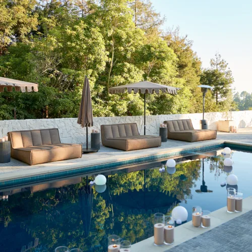 Poolside lounge area with brown cushioned sunbeds and beige umbrellas along a stone patio, candles placed along the pool edge, and trees in the background.