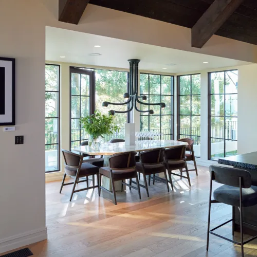 Bright dining area featuring a round marble table with brown chairs, a black multi-branch chandelier, and large windows letting in sunlight.