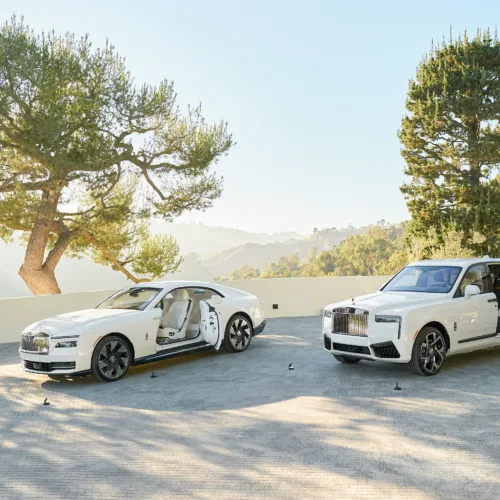 Two white luxury cars with doors open parked on a sunlit driveway, with trees and hills in the background.