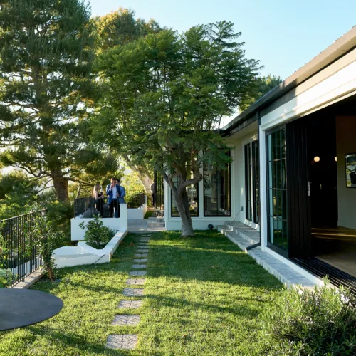 Modern house with large glass doors, a green lawn, and a stone path; a small group stands by a railing in the background.