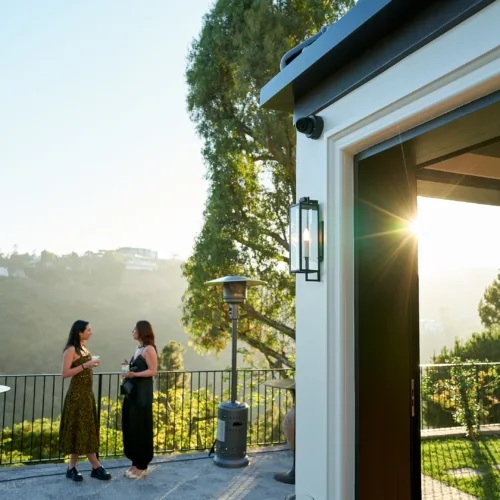 Two women in dresses chat on a sunlit outdoor patio with a railing and candle-lit glasses nearby.