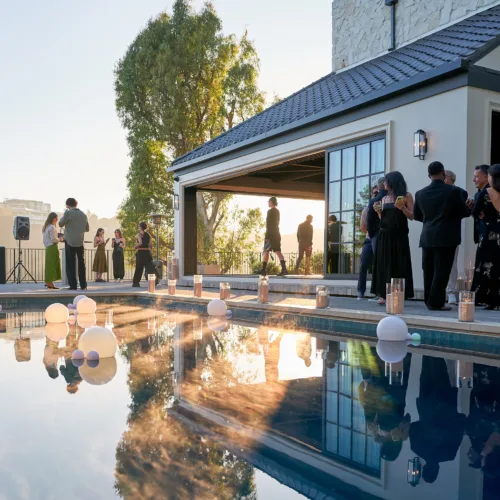 Outdoor evening party at a modern home with a pool; guests in formal attire mingle on the patio, balloons floating in the water.
