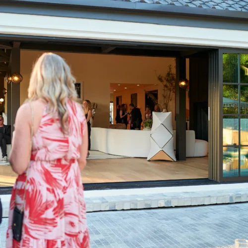 Guests in formal attire stand outside and inside a modern home during a party, with large sliding glass doors opened to a bright living area.