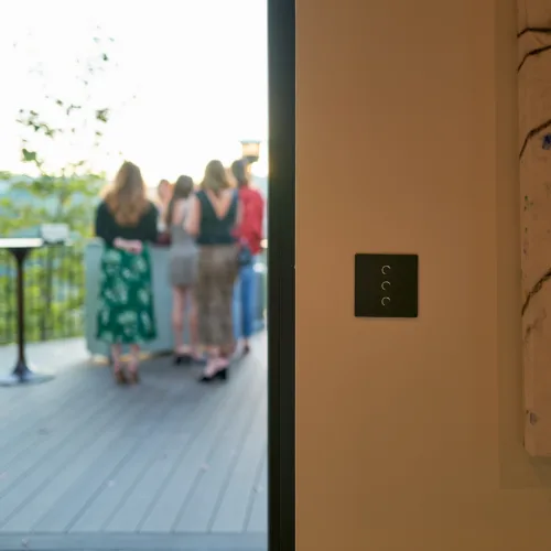 Group of people chatting on an outdoor deck seen through an open door, with a wall and light switch panel to the right.