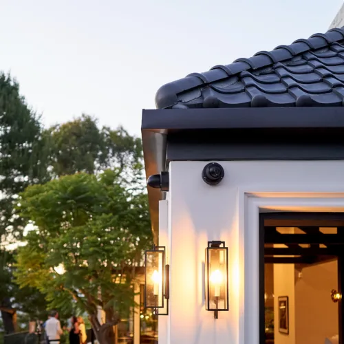 Open doorway at dusk with warm exterior lanterns, white frame, black-tiled roof, and people gathering in a garden background.