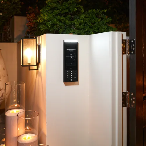 Electronic door intercom keypad mounted on a white wall pillar, with a green shrub above and lit glass candle holders nearby.