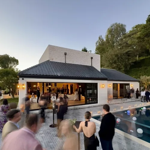 Outdoor evening party at a modern white house with a glass-walled terrace and a pool, guests mingling along the patio.